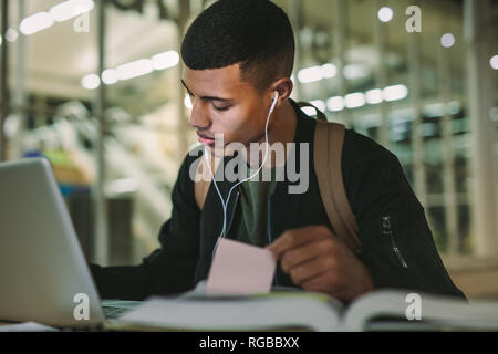 Male student sitting making notes with laptop on table preparing class assignments. Young man studying at college library. Stock Photo
