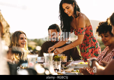 Beautiful woman serving food to friends sitting at dinner table. Group of friends having a party together outdoors. Stock Photo