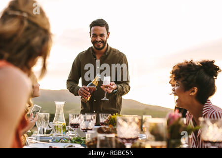 Smiling man serving champagne to friends sitting around table during party. Multi-ethnic group of friends having party outdoors. Stock Photo