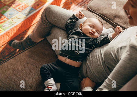 Baby in a deep sleep in the lap of his mother at home. Mother sitting on couch with her baby sleeping in her lap. Stock Photo