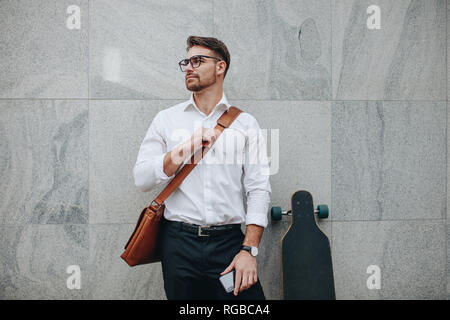 businessman wearing an office bag standing against a wall holding mobile phone. Formally dressed man standing with a long skateboard looking away. Stock Photo