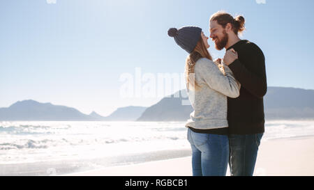Romantic couple standing at the sea shore. Man and woman standing close to each other on the beach looking at each other holding hands and smiling. Stock Photo