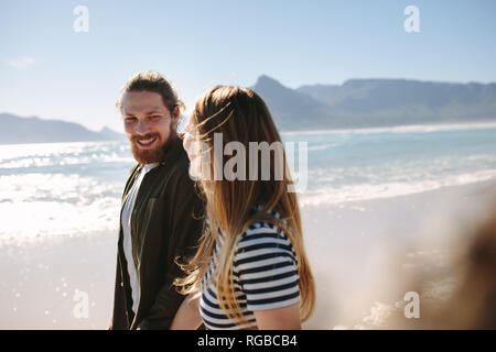 Handsome young man looking at his girlfriend while walking along the beach. Happy couple strolling outdoors on the beach. Stock Photo