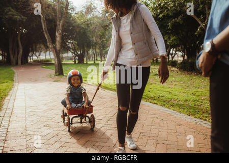 Mother pulling children in wagon at park. Boy wearing helmet sitting on a toy wagon being pulled by woman at park. Stock Photo