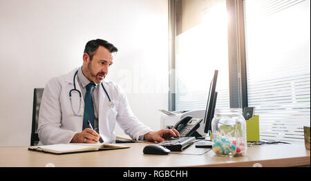 Doctor searching new information on internet using computer at his desk. Mature male medicine professional using computer and writing notes in diary. Stock Photo