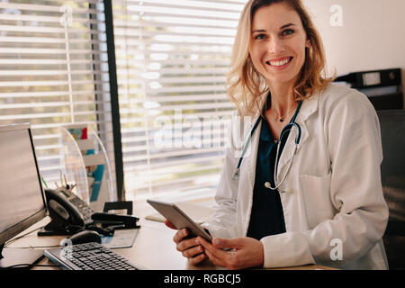 Female doctor sitting at her desk holding a digital tablet. Positive female physician looking at camera with smiling on the face. Stock Photo