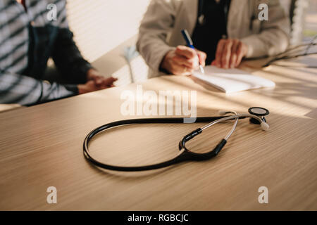 Stethoscope on table with female doctor prescribing drugs to her patient. Physician writing medicine prescription for the male patient. Stock Photo