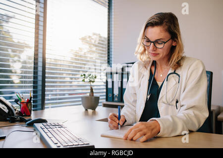 Female doctor sitting at the desk and writing prescription. Professional healthcare worker writing notes in a book. Stock Photo