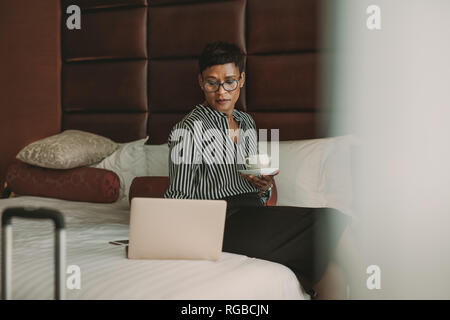 African woman in formal clothes sitting on bed with coffee in hotel room using laptop. Businesswoman working from hotel room. Stock Photo