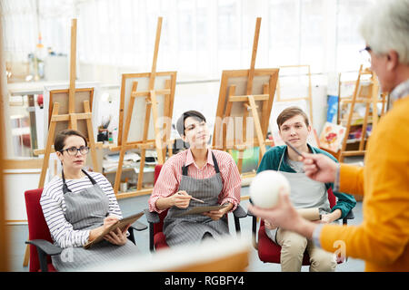 Portrait of group of cheerful art students sitting in row and listening to teacher during sketching session or lecture in class Stock Photo