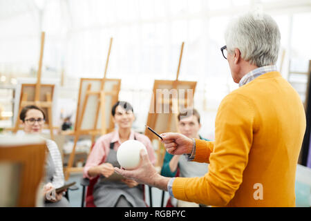 Rear view portrait of mature art teacher holding sphere and explaining light and shadow to group of students in art class, copy space Stock Photo