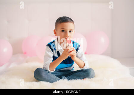 Portrait of white Caucasian cute adorable boy child eating heart shaped lollipop celebrating Valentine Day holiday. Kid eating candy sitting on bed in Stock Photo