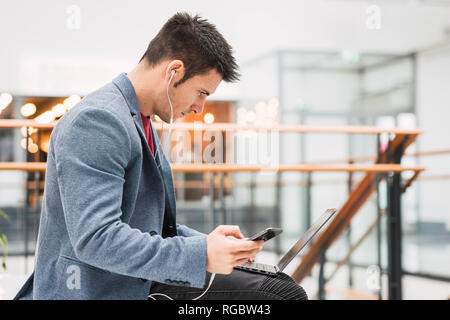 Businessman in lobby of a modern building, using smartphone and laptop Stock Photo