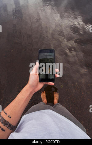 Man taking a cell phone picture with feet in water of a lake Stock Photo