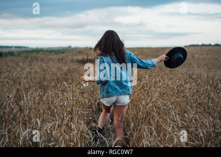 Back view of young woman with beverage and hat walking in corn field Stock Photo