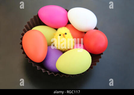 Yellow chick sitting with multi colored easter eggs in a brown basket against a dark grey background. Stock Photo