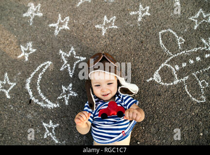 Portrait of smiling baby girl wearing pilot hat lying on asphalt painted with airplane, moon and stars Stock Photo