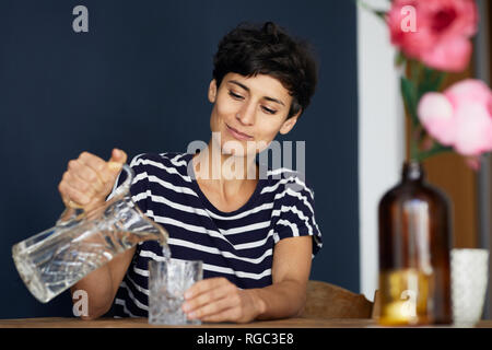 Woman at home sitting at wooden table pouring water into glass Stock Photo