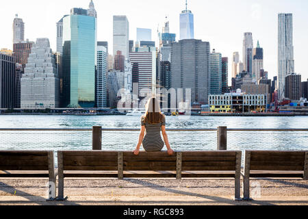 USA, New York, Brooklyn, back view of woman sitting on bench in front of East River and skyline of Manhattan Stock Photo