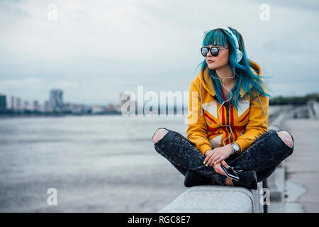 Young woman with dyed blue hair sitting on a wall listening music with headphones and smartphone Stock Photo