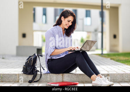 Smiling student sitting on stair outdoors working on laptop Stock Photo