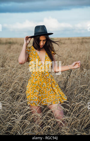 Young woman wearing summer dress with floral design and a hat dancing in corn field Stock Photo