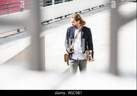 Young businessman carrying skateboard, using smartphone Stock Photo