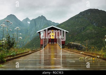 Man in yellow rain jacket waiting at red wood shack, Lapland, Norway Stock Photo