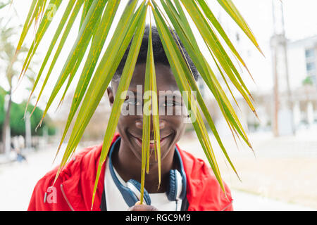 Young black man with headphones behind a palm leaf, smiling, portrait Stock Photo