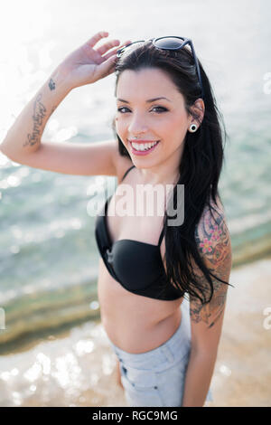Portrait of smiling young woman with nose piercing and tattoos standing at seashore