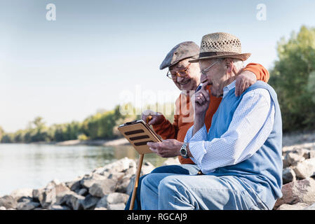 Two old friends sitting by the riverside, using digital tablet Stock Photo