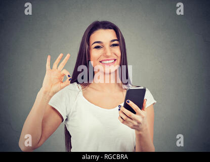Portrait of a smiling young cute woman holding blank screen mobile phone and showing ok sign over gray wall background Stock Photo