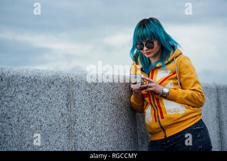 Portrait of young woman with dyed blue hair leaning against wall looking at cell phone Stock Photo