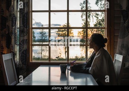 Finland, Lapland, young woman sitting at the window looking at a lake Stock Photo