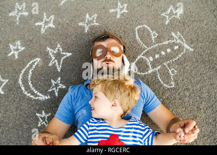 Portrait of mature man wearing pilot hat and his little son lying on asphalt painted with airplane, moon and stars Stock Photo