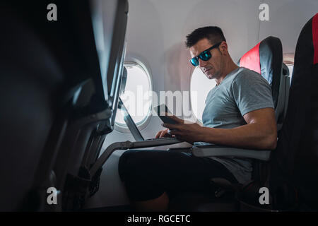 Man in airplane, using smartphone and laptop Stock Photo