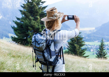 Germany, Bavaria, Oberammergau, young woman hiking taking a cell phone picture on mountain meadow Stock Photo