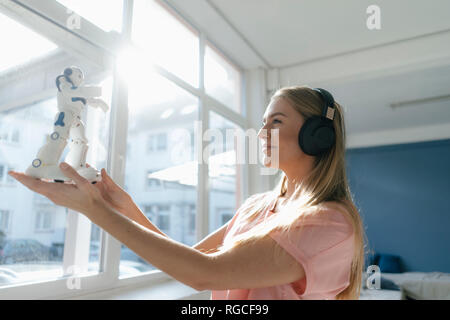 Smiling young woman with headphones looking at toy robot Stock Photo