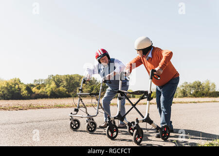 Two old friends wearing safety helmets, competing in a wheeled walker race Stock Photo