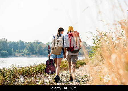 Rear view of young couple with backpacks and guitar walking at the riverside Stock Photo