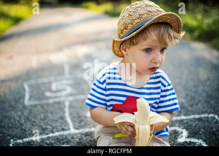 Portrait of toddler boy with banana sitting on the street watching something Stock Photo