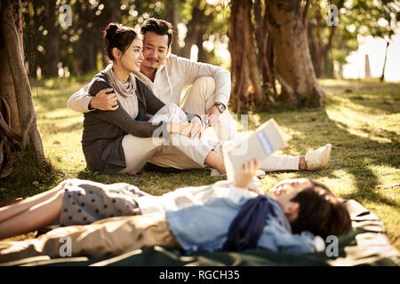 young asian couple sitting on grass in park chatting with two children lying reading book in foreground. Stock Photo