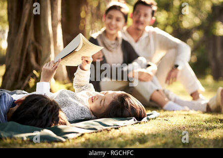 two asian children little boy and girl having fun lying on grass with parents sitting watching in background, focus on the children in foreground. Stock Photo