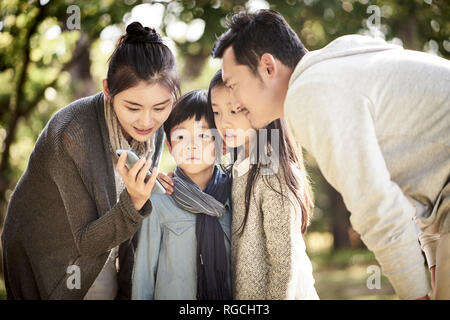 asian family parents and two children looking at mobile phone together in a park. Stock Photo
