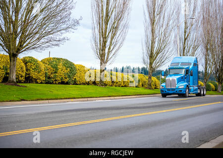 Classic bonnet commercial transportation pro American big rig blue long haul semi truck tractor driving on the road with trees and bushes for timely a Stock Photo