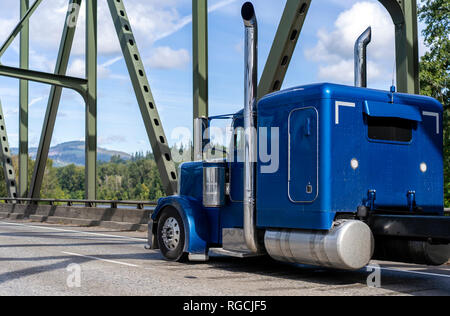 Bright blue classic American idol big rig bonnet semi truck tractor with driver rest compartment and high exhaust pipes running on the arched metal tr Stock Photo