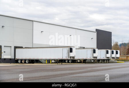 Long refrigerator semi trailers stand in row in warehouse docks with gates and loading and unloading commercial cargo and continuing go to the destina Stock Photo