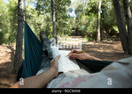 Man lying in hammock in the woods relaxing with puzzle book, partial view Stock Photo