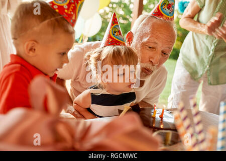 Grandfather and grandchildren blowing out candles on birthday cake on a garden party Stock Photo