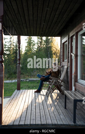 Young man sitting on veranda of a wood house, playing the ukulele Stock Photo